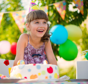 Little girl laughing at birthday party, wearing birthday hat and sitting in front of a cake with balloons in the background